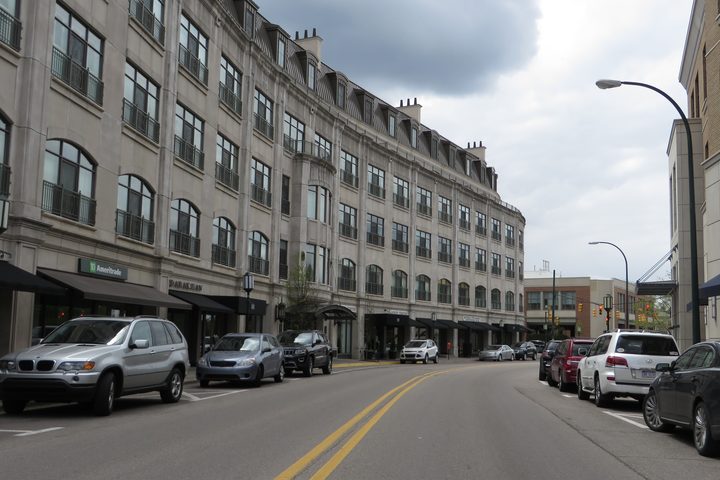 A four-story limestone building running down a Birmingham street: shops at ground level under awnings, office windows in the floors above them, and cars parked at an angle along the curb.