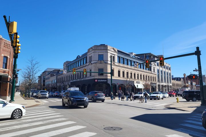 The corner of Maple Road and Old Woodward in downtown Birmingham, Michigan on a clear day: a limestone building on the far corner with storefronts beneath it, cars stopped at the light and people crossing.
