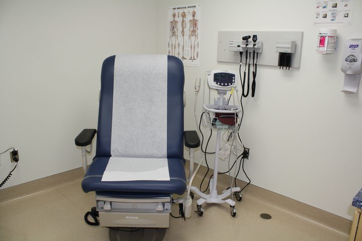 A clinical examination room: an upholstered exam chair with paper laid over it, a vitals monitor on a stand, a wall-mounted otoscope and a sharps box.
