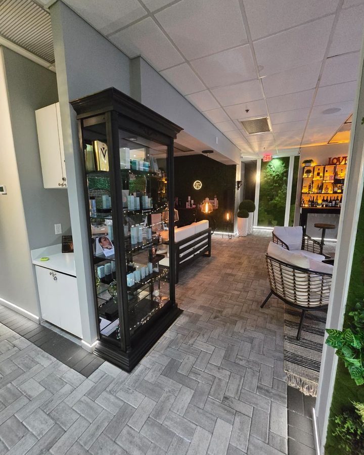The lounge of a med spa: a lit product cabinet against one wall, a herringbone floor, seating at the far end and planting by the window.