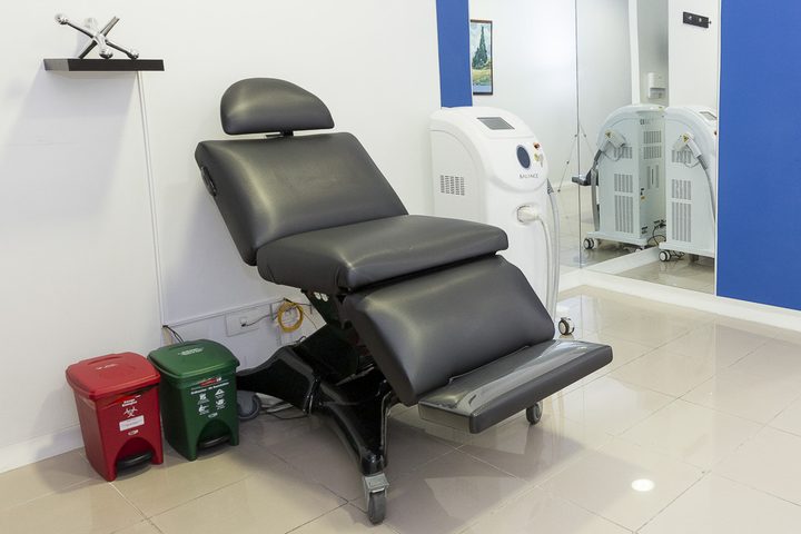 A bright treatment room with a reclining procedure chair on castors, a headrest, an instrument trolley beside it and cabinets along the far wall.