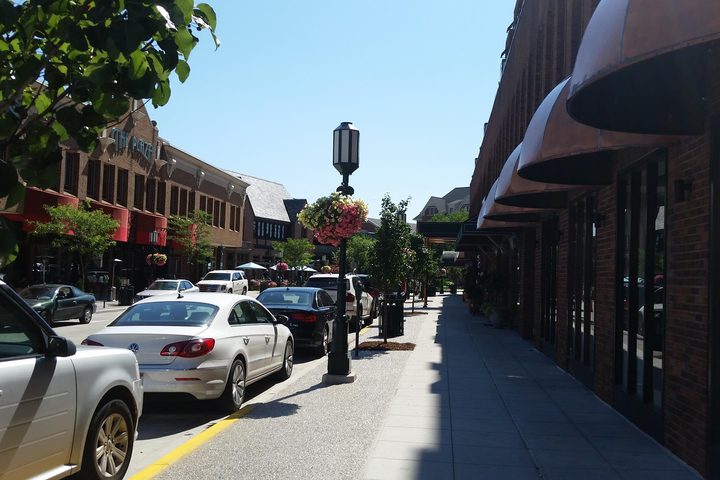 A Birmingham, Michigan sidewalk at street level: awnings running down the block, a cast-iron street lamp with a hanging flower basket, cars parked along the curb and tables further down the street.