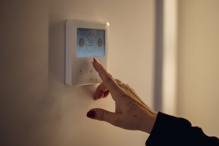 A hand reaching up to a wall thermostat in a hallway, the setpoint lit on its screen